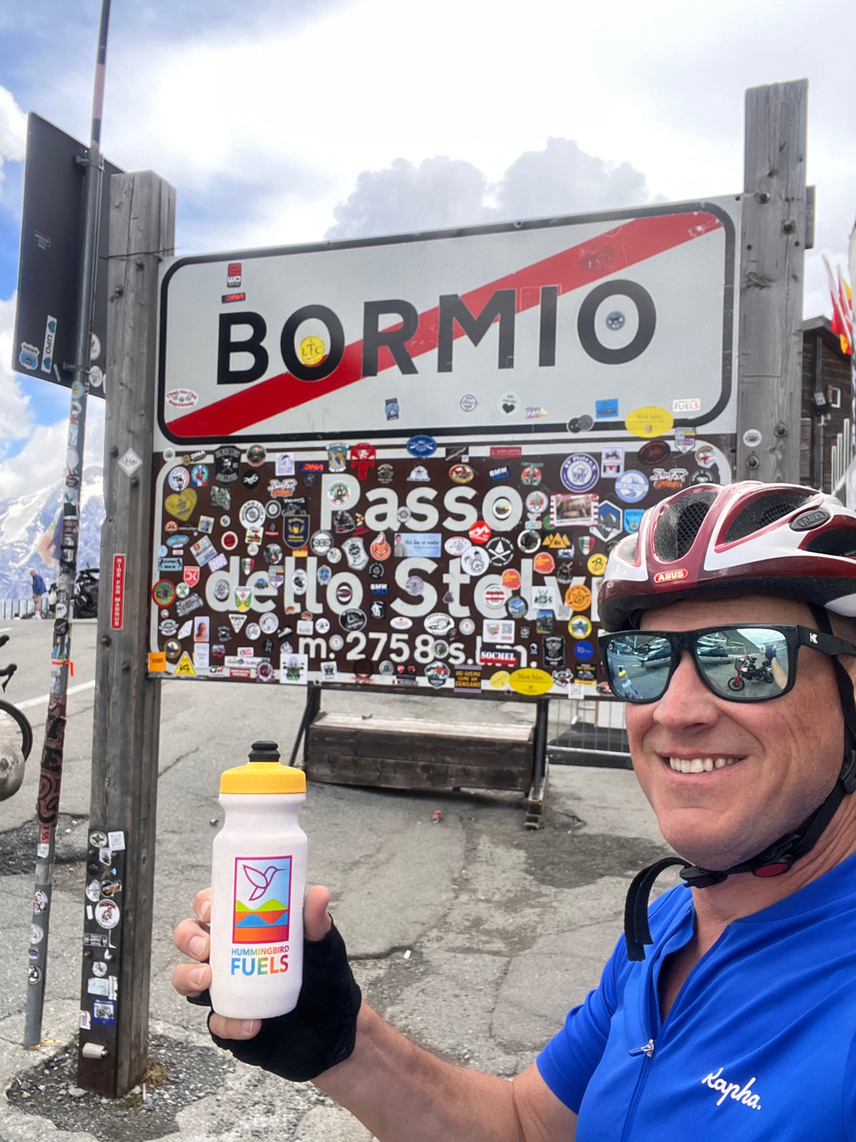 A smiling man in a blue Rapha cycling jersey, helmet, and sunglasses holds up a white Hummingbird Fuels bottle with a yellow cap. He stands in front of a large 'BORMIO' sign covered in stickers, with a mountain road and cloudy sky in the background.