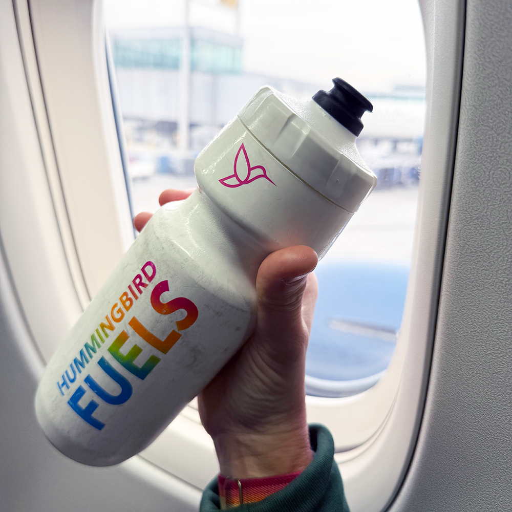 Hand holding a 'Hummingbird Fuels' bottle in front of an airplane window with a view of the runway.