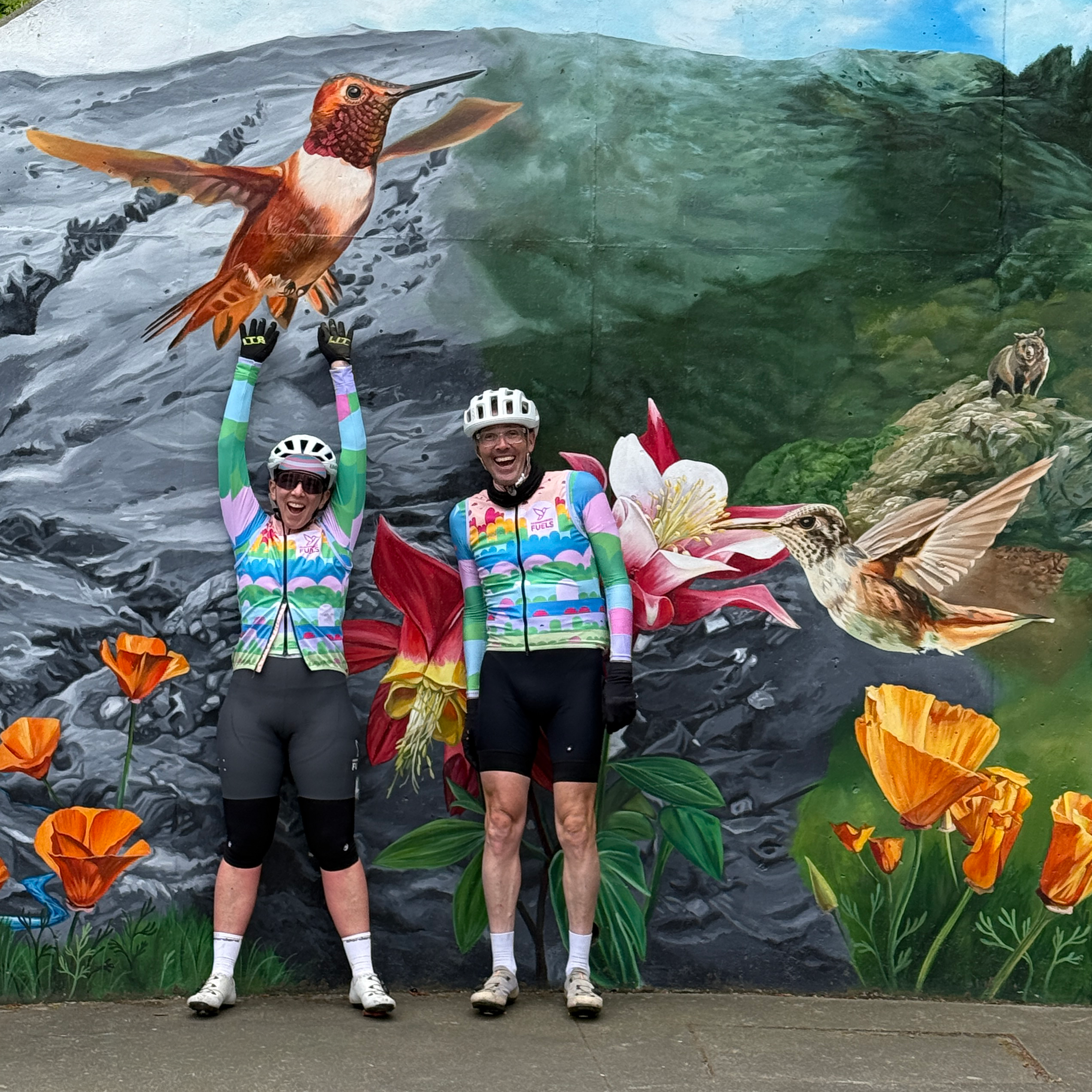 Two people in colorful cycling outfits posing in front of a mural with hummingbirds and flowers.