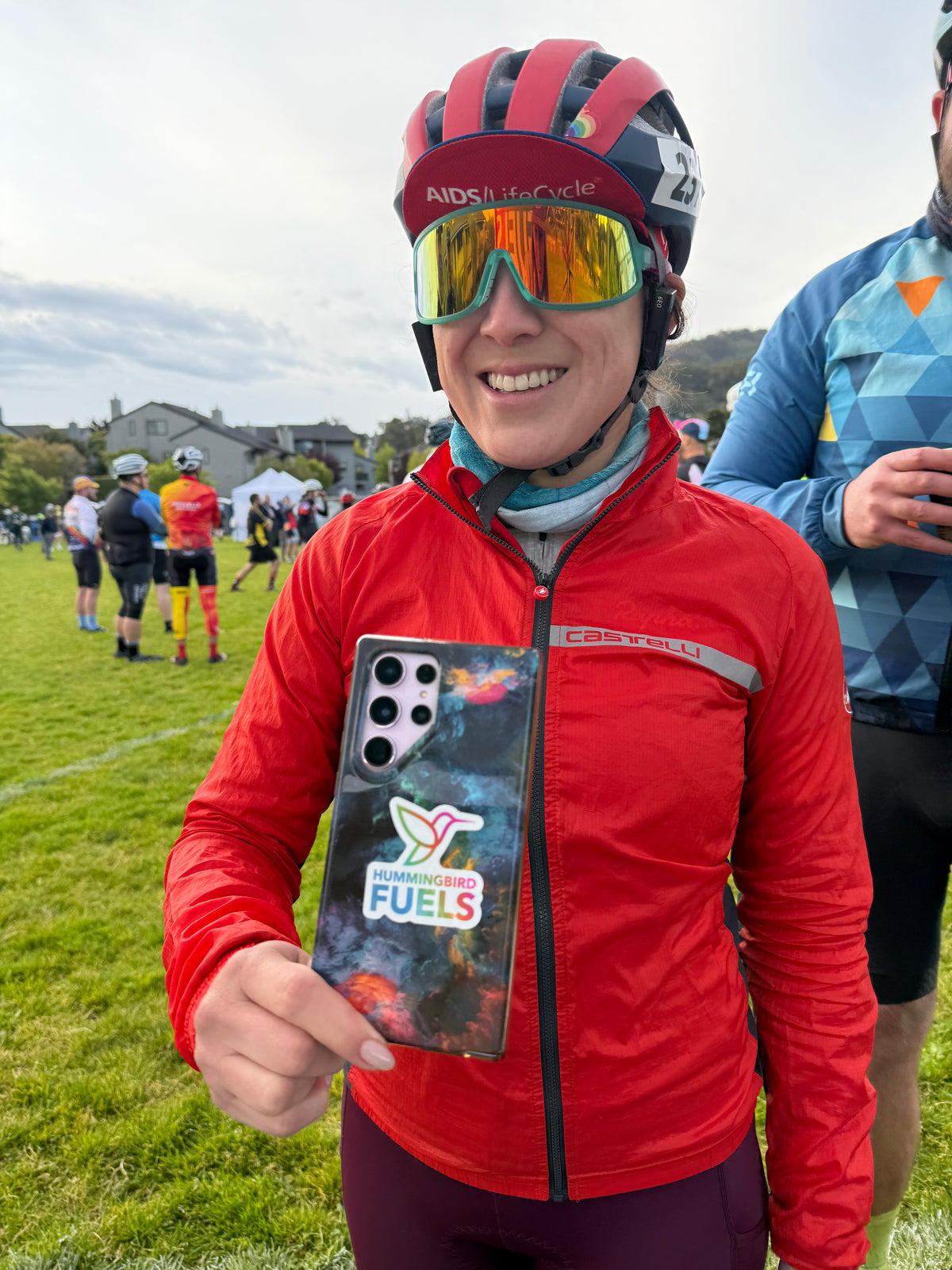 A smiling woman in a red cycling jacket and helmet holds up a custom phone case featuring a colorful galaxy design and a Hummingbird Fuels sticker. The background shows an outdoor grassy field with other cyclists and a cloudy sky.