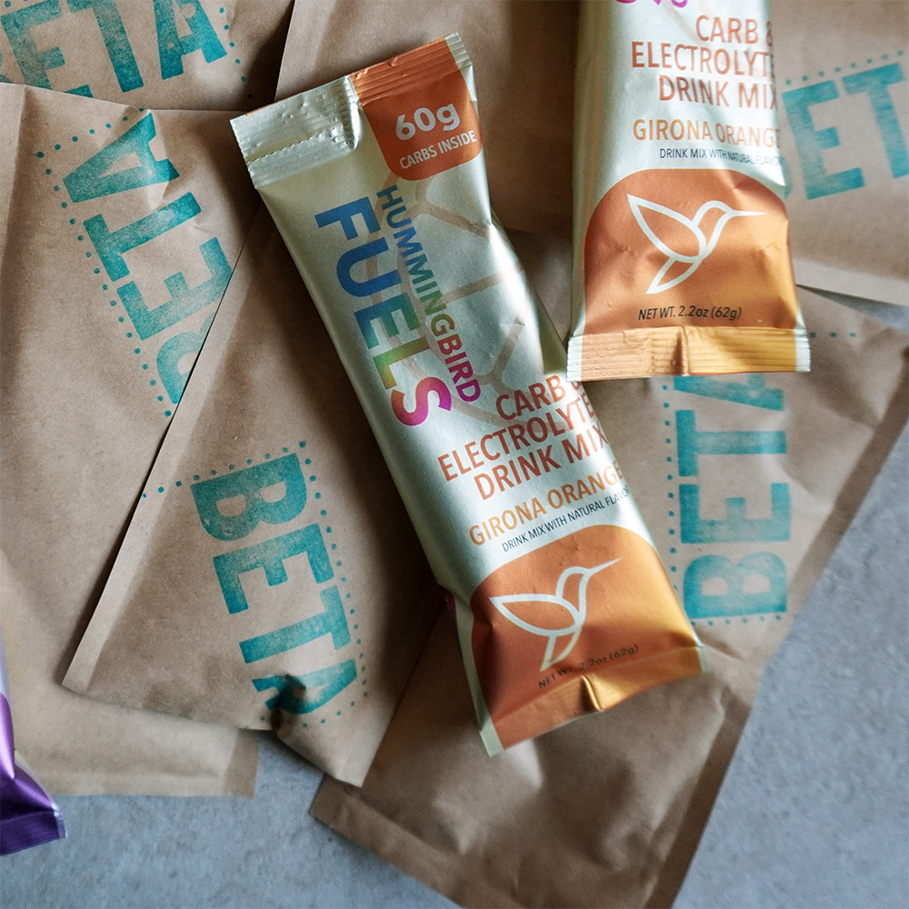 Hummingbird Fuels Magic Gel BETA packets with a representative Girona Orange single serve Sachet and container on a textured surface