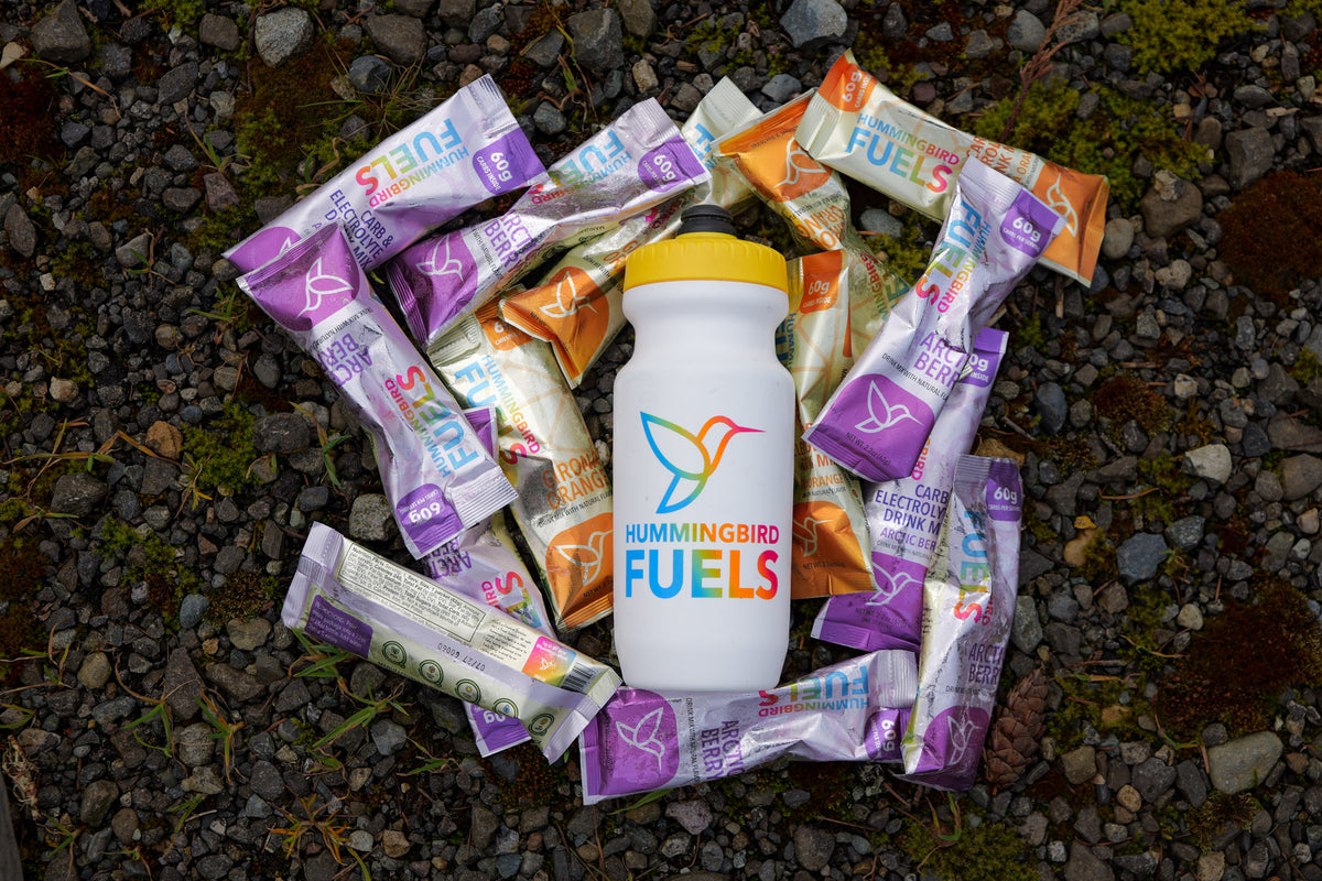 A white Hummingbird Fuels bottle with a yellow lid sits on a bed of dark gravel, surrounded by multiple single serve sachets