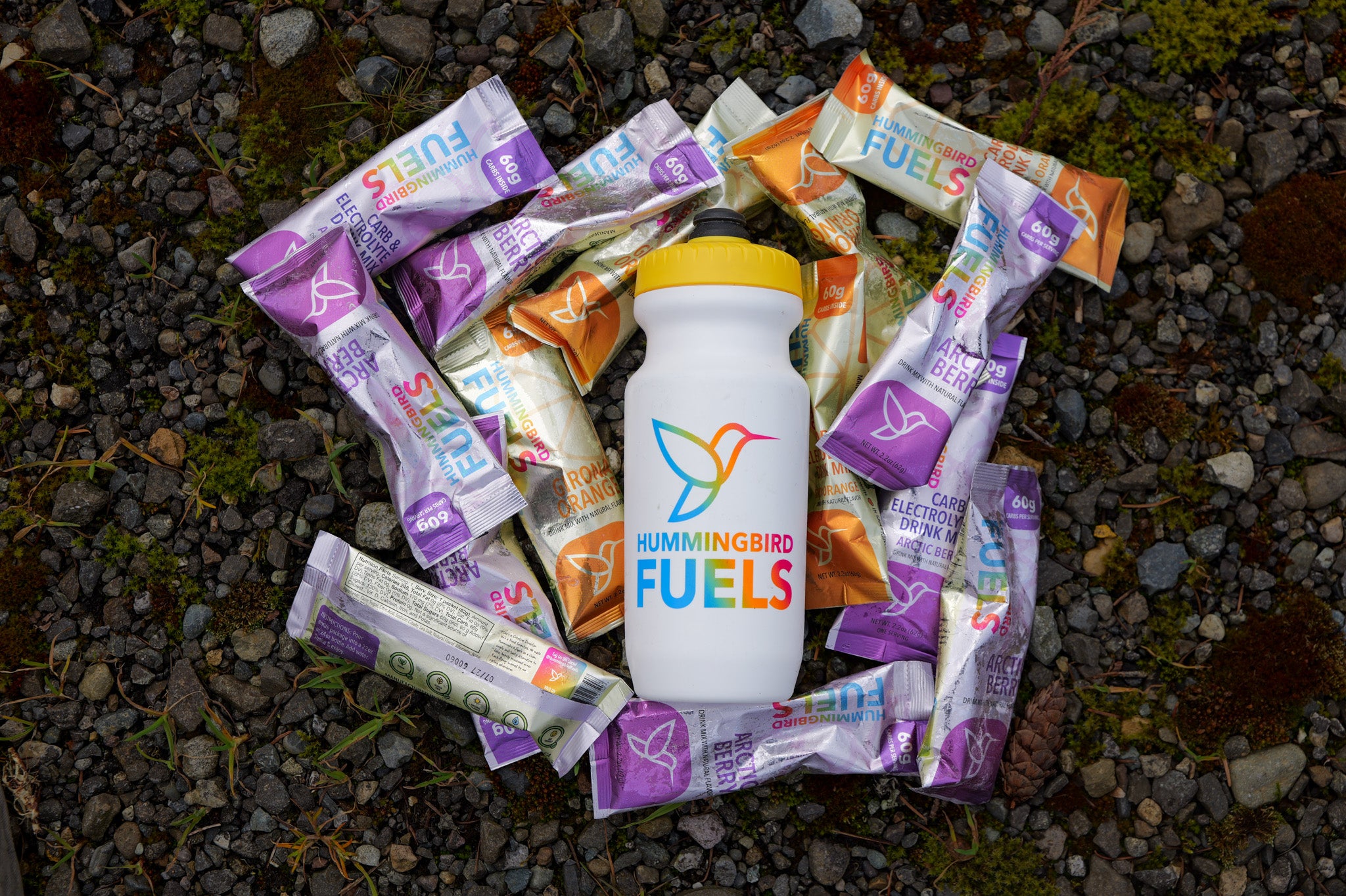 A white Hummingbird Fuels bottle with a yellow lid sits on a bed of dark gravel, surrounded by multiple single serve sachets